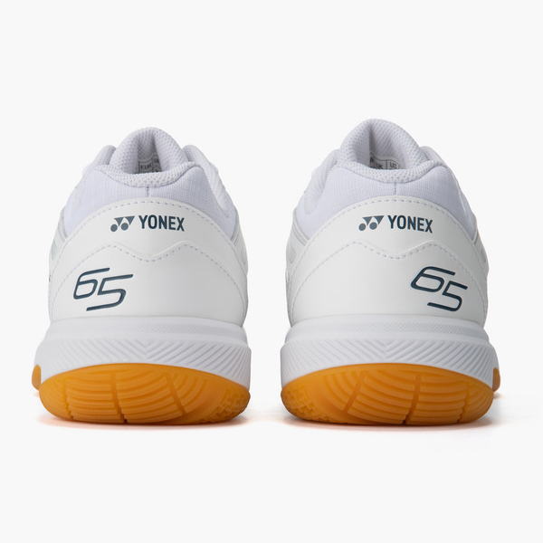 Pair of white Yonex tennis shoes viewed from the back, showing orange outsoles and blue '65' logos on the heel.